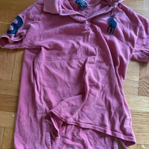 Rosa pikétröja från Ralph Lauren - Säljer en snygg rosa pikétröja från Ralph Lauren i bra skick. Den har korta ärmar och en klassisk krage. På bröstet finns det ikoniska broderade loggan och en stor siffra 3 på ärmen. Perfekt för en avslappnad men stilren look!