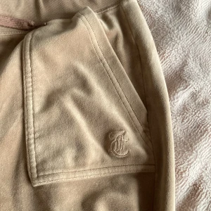Beige mjukisbyxor från Juicy Couture - Säljer ett par beige mjukisbyxor från Juicy Couture i storlek S. Byxorna har en bekväm passform med snörning i midjan och praktiska fickor fram och bak. Perfekta för en avslappnad stil.