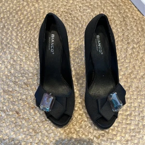 Svarta pumps från Bianco - Snygga svarta pumps från Bianco med en elegant klack och öppen tå. De har en stor dekorativ sten framtill som ger en lyxig touch. Perfekta för festliga tillfällen eller en utekväll. Storlek 37. Aldrig använda!