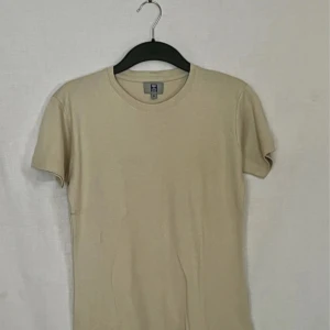 Beige t-shirt från Hallmans - Säljer en enkel och stilren beige t-shirt från Hallmans. Den är gjord i 100% bomull vilket gör den superbekväm och perfekt för vardagsbruk. T-shirten har en normal passform och korta ärmar, vilket gör den idealisk för sommaren. Perfekt basplagg att ha i garderoben!