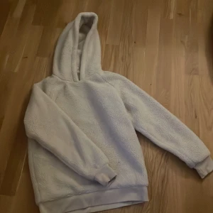 Vit teddy hoodie från Urban Classics - Mysig vit hoodie i teddy från Urban Classics. Perfekt för kyliga dagar med sin fluffiga och varma känsla. Den har en stor huva och långa ärmar med ribbade muddar. Passar perfekt för en avslappnad stil eller mysiga hemmakvällar.