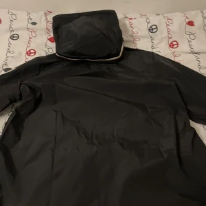 Monclear windbreaker - Säljer en snygg svart vindjacka från Moncler. Den har en dragkedja framtill och en justerbar huva med röd och vit rand. Jackan har långa ärmar och två fickor med knappar. Perfekt för höst och vår! 🖤
