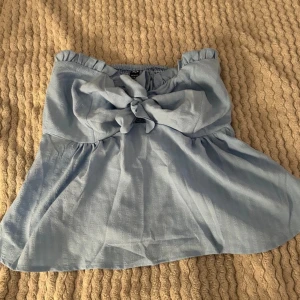 Ljusblå axelbandslös topp med rosett - Söt ljusblå axelbandslös topp med en stor rosett framtill. Perfekt för varma sommardagar och kan enkelt matchas med jeans eller kjol. Toppen har en luftig passform och är gjord i ett mjukt material.