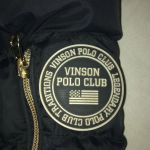 Svart dunjacka från Vinson Polo Club - Snygg svart dunjacka från Vinson Polo Club med pälskantad huva och dragkedja. Perfekt för kyliga dagar med sin varma och stilrena design. Säljer för att jag har köpt en ny jacka🌹