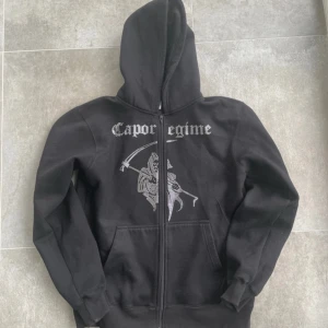 Svart hoodie från Costino Stockholm - Säljer en svart hoodie från Costino Stockholm med texten 'Caporegime' och en cool design på framsidan. Den har en dragkedja och är perfekt för höst och vinter. Grymt skön och stilren för alla tillfällen! Använd men i fint skick, inga defekter. Lite nopprig (se bild).