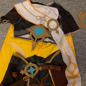 Genshin cosplay med peruk - Säljer en Aether cosplaydräkt med matchande peruk. Dräkten har en svart och gul design med vita och bruna detaljer, inklusive ett bälte. Peruken ingår i priset.