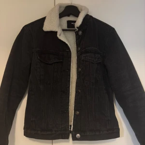Svart jeansjacka med teddyfoder från BikBok - Snygg svart jeansjacka från BikBok med mysigt vitt teddyfoder. Jackan har knappar framtill och två bröstfickor. Perfekt för kyligare dagar.