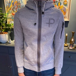 Grå hoodie från Pelle P - Snygg grå hoodie med dragkedja och justerbar huva från Pelle P. Tröjan har en praktisk ficka på ärmen.