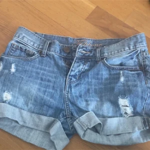 Blå jeansshorts från Liz Claiborne - Snygga blå jeansshorts från Liz Claiborne med slitna detaljer och uppvikta ben. Perfekta för en avslappnad sommarlook. De har en knapp och dragkedja framtill samt klassiska fem fickor.