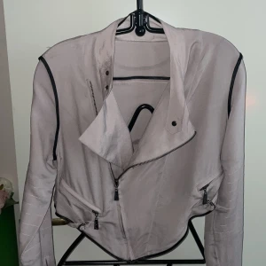 Gråbeige  jacka med dragkedja ny - Snygg gråbeige jacka med asymmetrisk dragkedja och svarta detaljer. Ny aldrig använt .Jackan har långa ärmar och flera dragkedjor framtill. Perfekt för en stilren look.