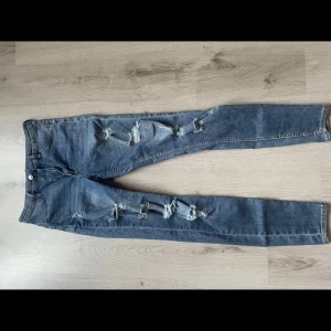 Blå slitna jeans - Säljer ett par blå jeans i bra skick med snygga slitningar längs benen. De har en klassisk femficksdesign och är perfekta för en avslappnad stil. Passar bra till både vardag och fest. Storlek 40.