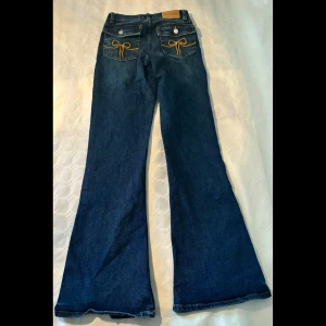 Blå jeans från Monki, bootcut och low waist - Säljer ett par snygga blå bootcut, low waist jeans från Monki. Broderad rosett på bakfickorna och knappar som detaljer. Oanvända. 
