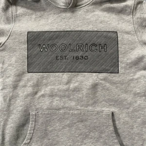 Grå hoodie från Woolrich - Säljer en snygg grå hoodie från Woolrich i storlek 14. Den har en stor ficka fram och ett stilrent tryck med 'Woolrich Est. 1830' på bröstet. Perfekt för kyliga dagar och ger en avslappnad look. 🩶För dig med stil woolrich,