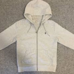 Grå hoodie från Polo Ralph Lauren - Säljer en snygg och bekväm grå hoodie från Polo Ralph Lauren. Den har en dragkedja framtill och en klassisk huva med dragsko. Perfekt för en avslappnad stil och passar till de flesta tillfällen. 🩶