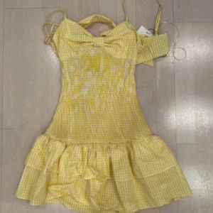 Gul rutig klänning med volang - gul klänning med rutigt mönster och volangdetaljer. Klänningen har en söt rosett framtill och är ärmlös med smala axelband. Perfekt för somriga dagar!