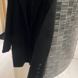 Svart kappa - Säljer en stilren svart kappa i bra skick. Den har en klassisk design med knappar framtill och långa ärmar. Perfekt för höst och vinter, och funkar både till vardags och fest. Passar bra över en tröja eller skjorta för en elegant look. S/M storlek