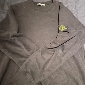 Svart sweatshirt från Stone Island - Säljer en svart sweatshirt från Stone Island i bra skick. Den har en klassisk rund hals och långa ärmar. På vänster ärm finns det ikoniska Stone Island-märket. Perfekt för höst och vinter, både stilren och bekväm.