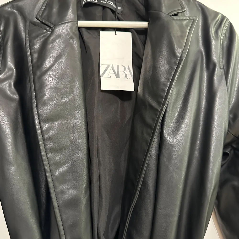 Snygg svart skinnjacka från Zara med knytbälte i midjan. Jackan har en klassisk krage och långa ärmar, perfekt för en stilren look. Går också att använda som en klänning. Aldrig använd säljs för 300kr . Takit.