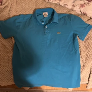 Blå pikétröja från Lacoste - Säljer en snygg blå pikétröja från Lacoste med klassisk krage och knappdetalj framtill. Tröjan har korta ärmar och den ikoniska krokodilloggan på bröstet. Perfekt för en stilren och avslappnad look