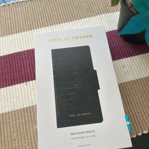 Svart mobilfodral från Ideal of Sweden - Säljer ett snyggt svart mobilfodral från Ideal of Sweden i modellen Neo Noir Croco. Passar iPhone 12/12 Pro. Fodralet har en stilren krokodilmönstrad design och är perfekt för att ge din mobil en lyxig look. Kommer i originalförpackning.