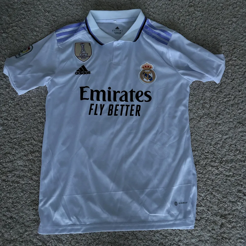 Säljer en vit Real Madrid fotbollströja från Adidas med korta ärmar. Tröjan har klubbmärket och 'Emirates Fly Better' tryckt på framsidan. På baksidan står det 'VINI JR.' och nummer 20. Den har även FIFA World Champions 2023-märket på bröstet. Perfekt för alla Real Madrid-fans!. T-shirts.