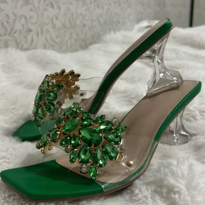 Gröna klackskor med strassdetaljer - Snygga gröna klackskor med transparent klack och glittriga strassdetaljer framtill. Perfekta för en festlig kväll eller bal. De har en fyrkantig tå och en unik design som verkligen sticker ut. Klacken är i en cool transparent stil som ger en modern touch.