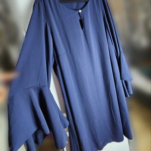 Mörkblå lång blus från Evans - Säljer en elegant mörkblå blus från Evans i storlek 52. Den har långa, vida ärmar med volangdetaljer och en fin pärlknapp vid halsen. Perfekt för höst- och vinterfester. Blusen är i nyskick och har ett robust tyg. 