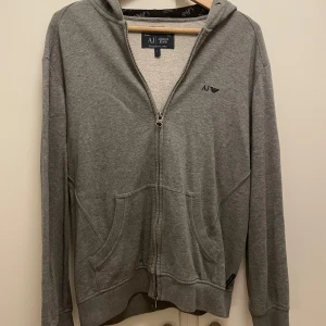 Grå hoodie från Armani Jeans - Säljer en snygg grå hoodie från Armani Jeans i storlek S. Den har en dragkedja framtill och en broderad logga på bröstet. Perfekt för en avslappnad stil och passar till alla årstider. Huvan är fodrad med ett mönstrat tyg för extra stilpoäng. 🩶
