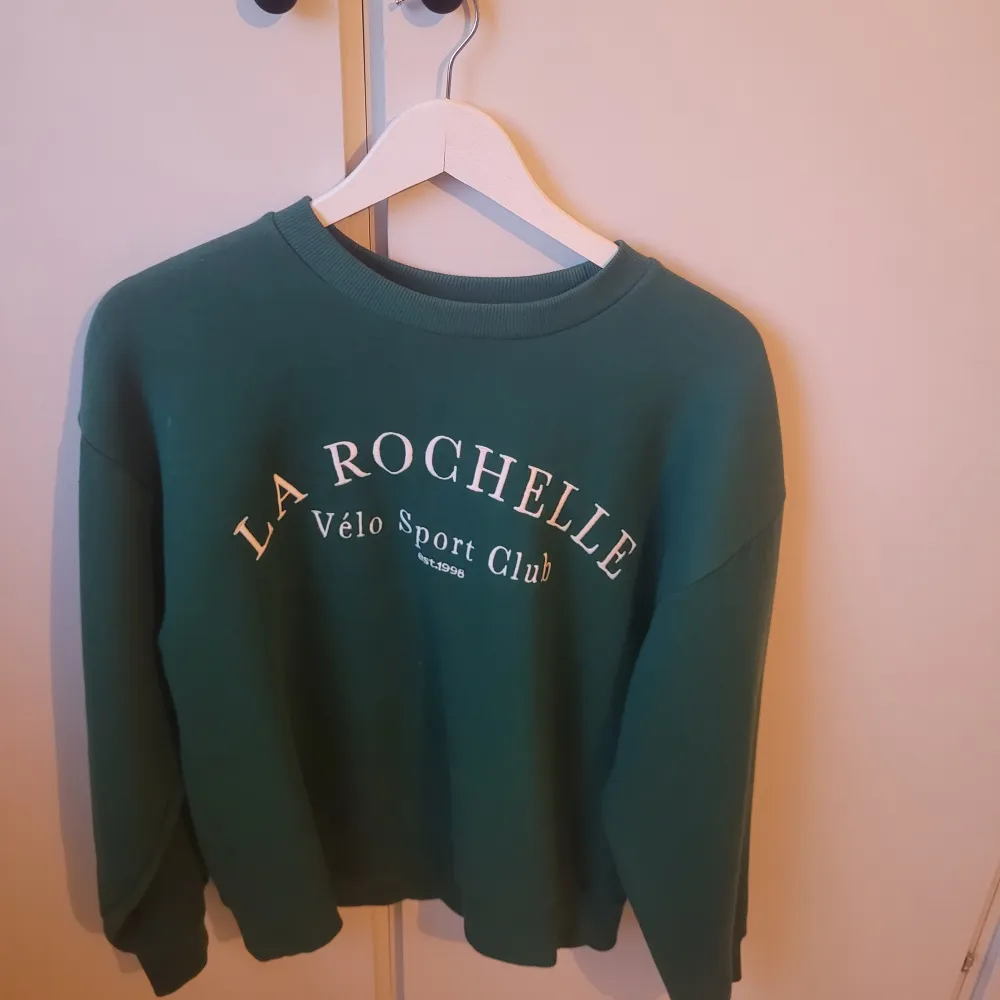 Säljer en snygg grön sweatshirt från Gina Tricot med texten 'La Rochelle Vélo Sport Club' tryckt på framsidan. Tröjan har en klassisk rund hals och långa ärmar, perfekt för en avslappnad stil. Den är i mycket bra skick och passar perfekt för höst och vår.. Tröjor & Koftor.