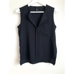 Svart enkelt linne, 34 XS, basic, stilren, enfärgad, topp, ärmlös - Svart linne i vävt material, en bröstficka, längre bak. Storlek (EU): 34, Bröstvidd: 44 cm, Axelbredd: 35 cm, Längd fram från axeln: 56,5 cm