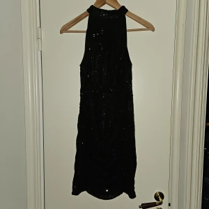 Svart paljettklänning nytt - Ny Snygg svart klänning med paljetter. Klänningen är figursydd och har en hög hals. Resår i midjan och dragkedja bak. Längd 86cn , bredd 37 cm Den är perfekt för festliga tillfällen.