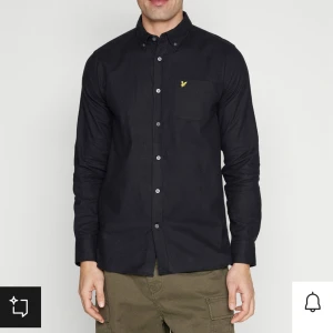 Svart skjorta från Lyle & Scott - Snygg svart skjorta från Lyle & Scott i storlek S. Den har långa ärmar och en klassisk krage. Skjortan är dekorerad med det ikoniska gula logotypen på bröstet. Perfekt för både vardag och fest!