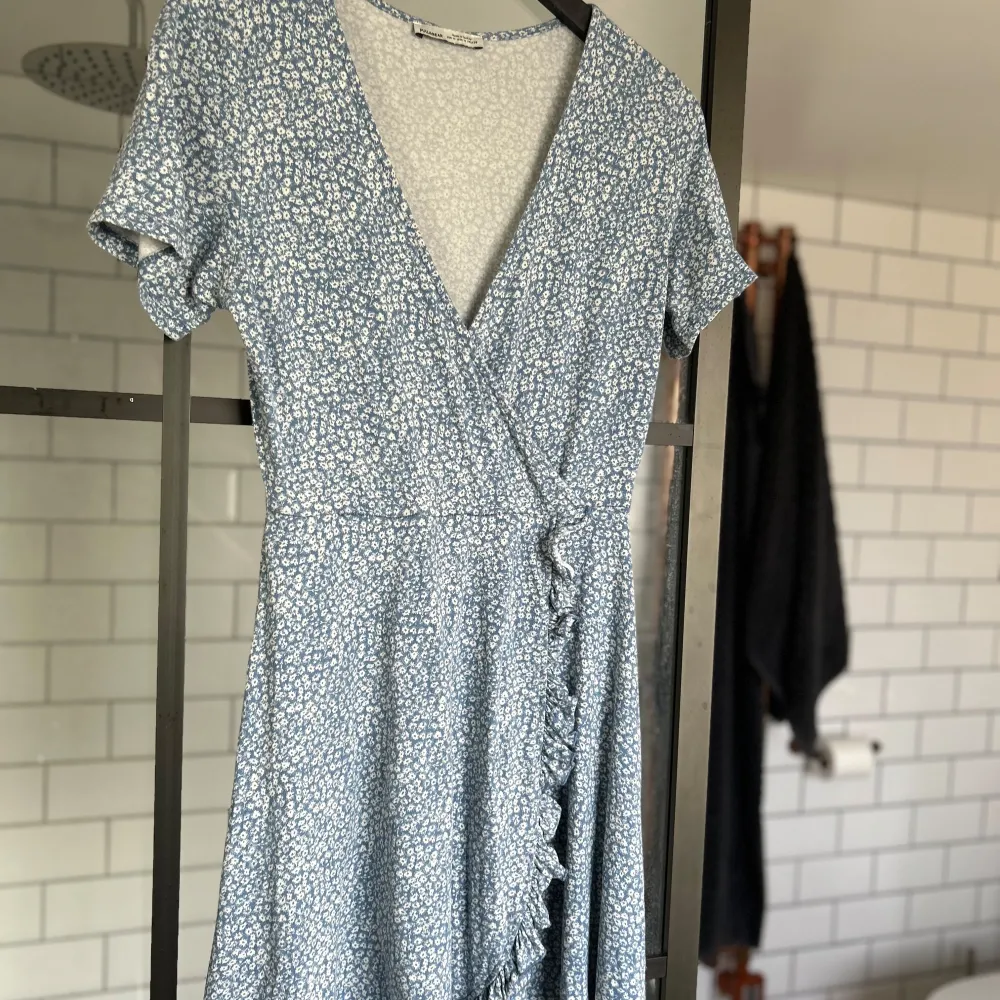 Säljer en superfin blå omlottklänning från Pull&Bear med ett sött blommönster. Klänningen har korta ärmar och en volangdetalj längs omlottkanten. Perfekt för vår och sommar! 🌸. Klänningar.