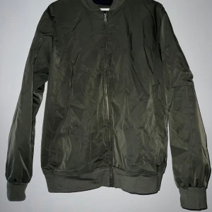Olivgrön bomberjacka - Säljer en snygg olivgrön bomberjacka i mycket bra skick. Jackan har en dragkedja framtill och långa ärmar med ribbade muddar. Perfekt för höst och vår, och den ger en cool look till vilken outfit som helst. Passar både till vardags och fest! Just nu är den lite skrynklig då den legat i en påse. Se sista bild för en liten defekt. Materialet är något skimmrigt.