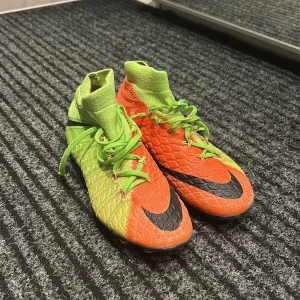 Färgglada fotbollsskor från Nike - Säljer ett par coola fotbollsskor från Nike i neonfärger. Skorna är i grönt och orange med svarta detaljer och har snörning. Perfekta för fotbollsträningen med bra grepp tack vare dobbarna. De är i mycket bra skick och redo för nya äventyr på planen!