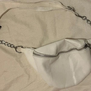 Vit axelväska med kedjedetaljer - Snygg vit axelväska i syntetmaterial med en cool kedja som axelrem. Väskan har en praktisk dragkedja upptill och är perfekt för både vardag och fest. Den är i bra skick och rymmer det viktigaste du behöver ha med dig. En stilren och modern accessoar för alla tillfällen! Säljs för 40