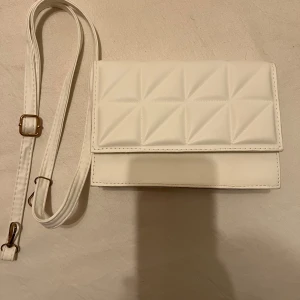 Vit quiltad axelväska - Snygg vit axelväska med quiltad design och justerbar rem. Perfekt för en stilren look, med magnetknappar för enkel öppning och stängning. Väskan är i syntetmaterial och passar perfekt till både vardag och fest. Säljs för 40