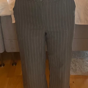 Grå randiga kostymbyxor - Säljer ett par grå kostymbyxor med vita ränder. De har en klassisk och stilren look, perfekt för både kontoret och mer formella tillfällen. Byxorna har en normal passform och är högmidjade för en bekväm och smickrande siluett. Det finns en liten fläck på ena benet, men annars i bra skick.