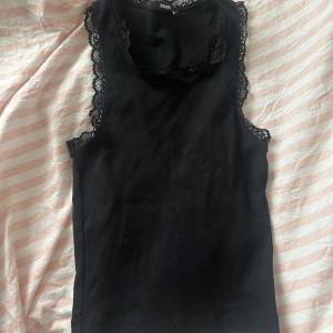 Svart ärmlös topp med spetsdetaljer - Snygg svart ärmlös topp från Bik Bok med fina spetsdetaljer runt ärmhålen och halsringningen. Perfekt för både vardag och fest! Toppen är i ett mjukt och bekvämt material som sitter skönt mot kroppen.