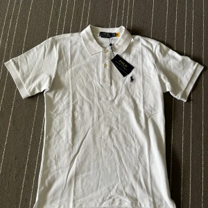 Vit pikétröja från Polo Ralph Lauren - Säljer en klassisk vit pikétröja från Polo Ralph Lauren i nytt skick. Tröjan har korta ärmar och en liten broderad logga på bröstet. Perfekt för en stilren och avslappnad look. Passar både till vardags och fest! 🕶️