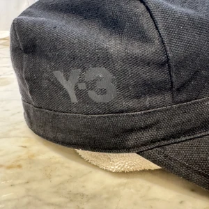 Svart keps från Y-3 - Säljer en stilren svart keps från Y-3, designad av Yohji Yamamoto. Kepsen är i storlek M och har en diskret logga på sidan. Perfekt för både vardag och träning. Tillverkad i en blandning av linne och bomull för en bekväm passform.