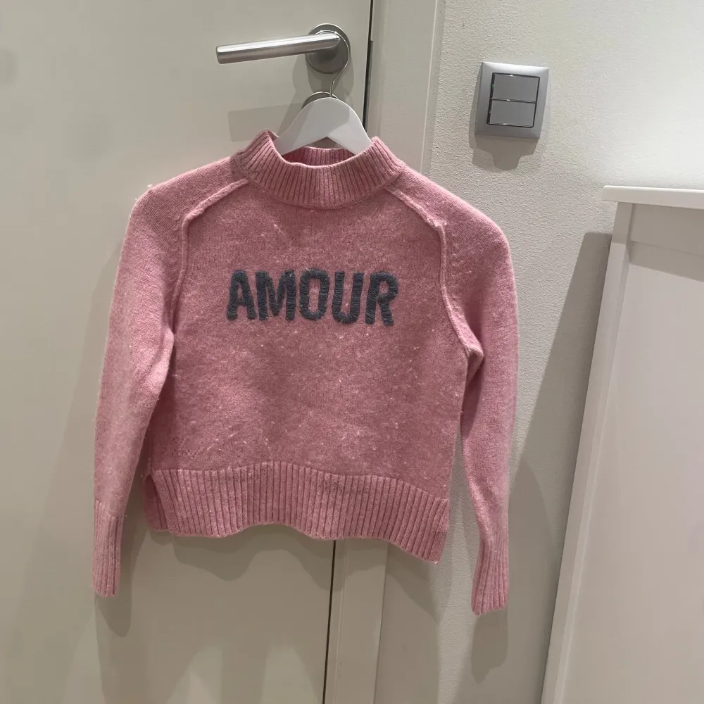 Superfin rosa stickad tröja från Zadig & Voltaire med texten 'AMOUR' i blåt på framsidan. Den har långa ärmar, perfekt för kyliga dagar. Tröjan är cool och har ribbade detaljer vid ärmslut och nederkant. Perfekt för en mysig höstl. Tröjor & Koftor.