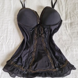 Svart korsett med spetsdetaljer - Snygg svart korsettopp med vackra gulddetaljer och små rosetter. Den har vadderade kupor och justerbara axelband för perfekt passform. Materialet är mjukt och glansigt, vilket ger en lyxig känsla. Perfekt för en festlig kväll.