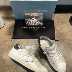  Philippe Model - Säljer ett par snygga vita sneakers från Philippe Model. De har en stilren design med vita snören och en kontrasterande mörkblå detalj vid hälen. Skorna är i skinn och har en klassisk sneakerstil som passar perfekt till både vardag och fest. De är i bra skick med lite slitage på sulorna.