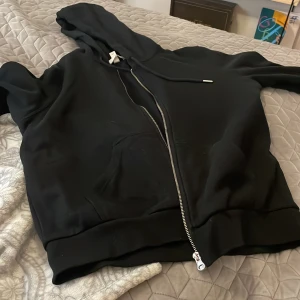 Svart hoodie med dragkedja - Säljer en svart hoodie i storlek M. Den har en dragkedja framtill och en mysig huva med snören. Perfekt för en avslappnad stil och passar till alla tillfällen. Skön och enkel att matcha med andra plagg!