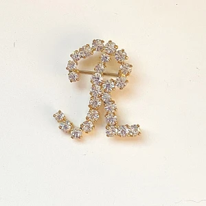 Guldfärgad brosch med strass - Säljer en superfin vintage guldfärgad brosch formad som bokstaven 'R'. Den är täckt med glittrande strass som verkligen fångar ljuset. Perfekt för att piffa upp en outfit till fest eller speciella tillfällen. Broschen är ungefär 4 cm lång. Okänd meta