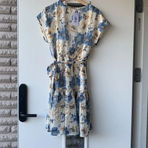 Blommig klänning från Ellos - Säljer en superfin blommig klänning från Ellos i storlek 38/40. Den har ett vackert blommönster i blått och beige på en ljus bakgrund. Klänningen är kortärmad och har en knytning i midjan för en smickrande passform. Perfekt för sommarens alla fester eller en dag på stan!