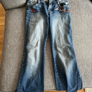 Blå jeans med broderade detaljer - Säljer ett par snygga blå jeans från EDC med coola broderade detaljer i rött och blått. Jeansen har en straight passform och är perfekta för en avslappnad stil. De har en klassisk femficksdesign och är i bra skick. Perfekta för både vardag och fest!