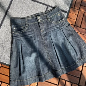 Jeanskjol med veck - Säljer en snygg jeanskjol i mörk denim med stilrena veck framtill. Kjolen har en klassisk femficksdesign och stängs med två knappar och dragkedja. Perfekt för en casual look och passar till både vardag och fest. Den är i bra skick och redo för en ny ägare!