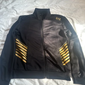 Svart jacka från Emporio Armani - Säljer en snygg svart jacka från Emporio Armani med coola guldfärgade detaljer. Jackan har en dragkedja framtill och EA7-loggan på bröstet. Perfekt för en stilren look!